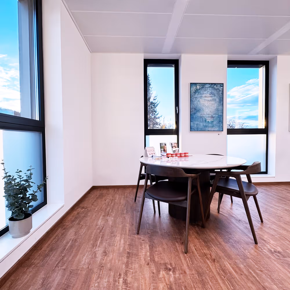 Moderner Besprechungsraum mit rundem Tisch, vier St&uuml;hlen, gro&szlig;en Fenstern und einem kleinen Topf mit Pflanze am Fenster.