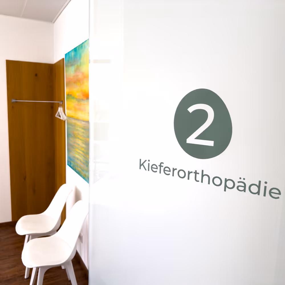 Behandlungsraum mit zwei wei&szlig;en St&uuml;hlen und einem bunten Gem&auml;lde an der Wand dahinter.