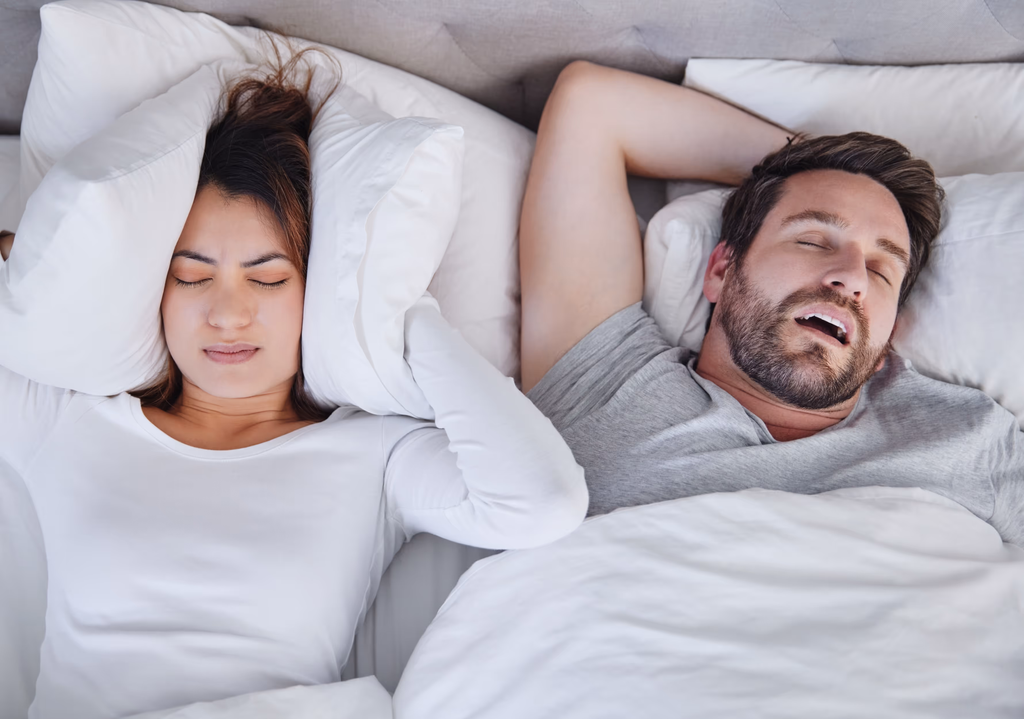 Frau im Bett h&auml;lt Kissen an die Ohren, w&auml;hrend neben ihr ein Mann mit offenem Mund schl&auml;ft.
