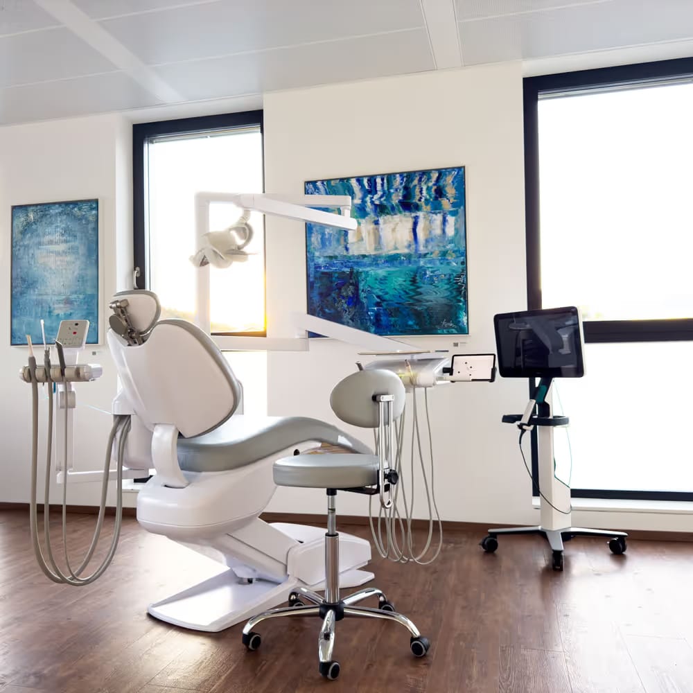 Heller Behandlungsraum mit einem weißen Zahnarztstuhl, Instrumenten und modernem 3D-Scanner vor großen Fenstern.