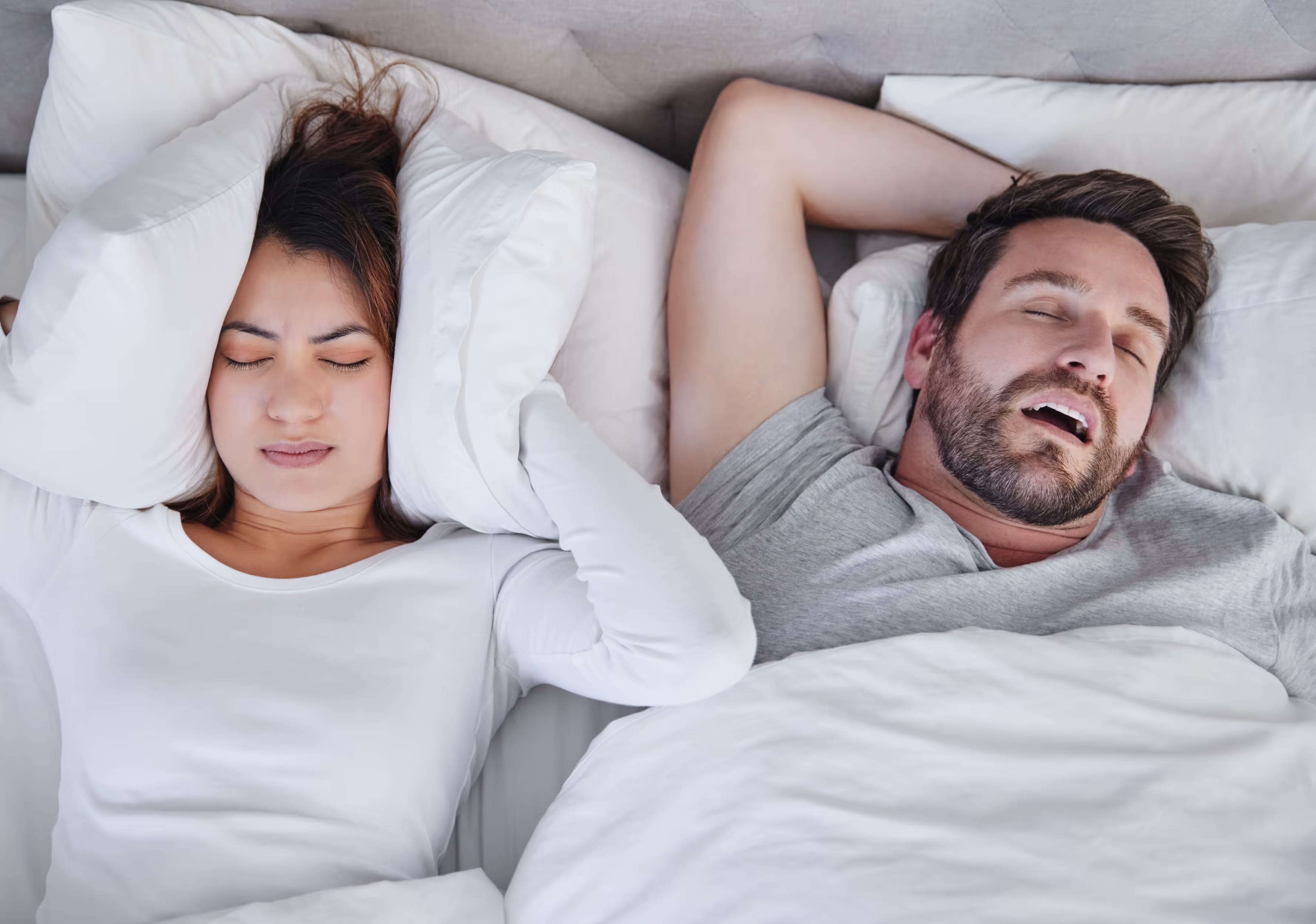 Frau im Bett hält Kissen an die Ohren, während neben ihr ein Mann mit offenem Mund schläft.